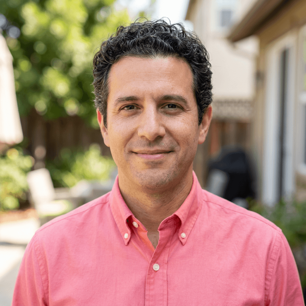 Smiling man with dark curly hair wearing a pink button-down shirt in a sunny backyard.